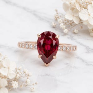 Könnte beinhalten: Ein Ring aus Roségold mit einem großen, birnenförmigen, tiefroten Edelstein, der von vier Krappen gehalten wird. Die Ringschiene ist mit einer Reihe kleiner, runder, klarer weißer Diamanten besetzt, die beidseitig des Mittelsteins funkeln.