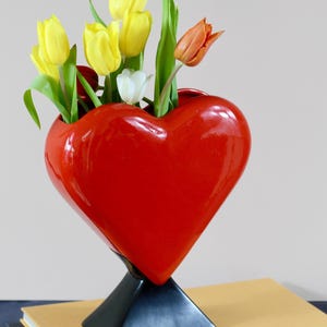 Op de afbeelding: Een glanzende, hartvormige rode vaas met gele, witte en oranje tulpen. De vaas staat op een zwarte geometrische basis. De bloemen hebben groene stelen en bladeren. De vaas staat op een stapel boeken.