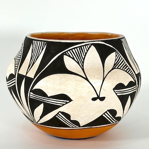 Op de afbeelding: Een handgemaakte keramische kom met een afgeronde vorm. De kom heeft een zwart-wit geometrisch ontwerp met een bloemmotief. De rand en de basis zijn geschilderd in tinten oranje en bruin.