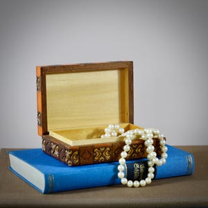 Könnte beinhalten: Eine offene Holzschmuckschatulle mit aufwendigen Schnitzereien und orangefarbenen Akzenten, die auf einem blauen Buch liegt. Eine Kette aus weißen Perlen liegt daneben. Die Schatulle ist leer und bereit, Schätze aufzubewahren.