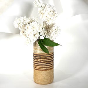 Könnte beinhalten: Eine zylindrische Keramikvase mit horizontalen braunen Streifen und einem beigefarbenen Sockel hält einen Strauß weißer Blumen und grüne Blätter. Die Vase hat eine strukturierte Oberfläche.