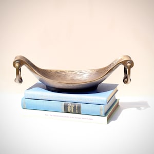 Könnte beinhalten: Eine bronzefarbene dekorative Schale mit gebogenen Griffen, die auf einem Stapel von drei blauen Büchern ruht. Die Schale hat eine strukturierte Oberfläche und eine bootsartige Form. Die Bücher haben einen hellblauen Einband.