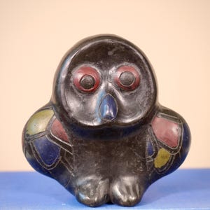 Könnte beinhalten: Eine dunkle, stilisierte Eulenfigur mit roten Augen und einem blauen Schnabel. Die Flügel der Eule sind mit farbenfrohen geometrischen Formen in Rot, Blau und Gelb verziert. Die Eule sitzt auf einer blauen Oberfläche.