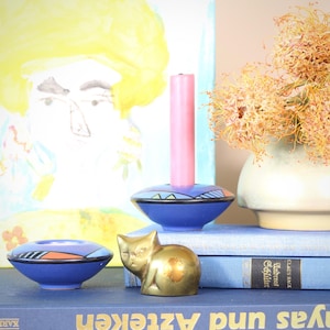 Può includere: Una natura morta con una candela rosa in un portacandele blu, una piccola ciotola blu e una statuetta di gatto in ottone. Un vaso con fiori secchi e un dipinto colorato sono sullo sfondo.