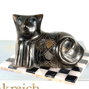 Könnte beinhalten: Eine schwarze Katzenfigur mit blauen Augen und aufwendigen Ätzdetails. Die Katze liegt auf einer karierten schwarz-weißen Marmorbasis. Die Figur hat ein einzigartiges, künstlerisches Design.