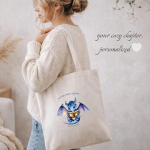 Könnte beinhalten: Cremefarbene Tragetasche mit einer Aquarellillustration eines blauen Drachen, der ein Buch liest. Auf der Tasche steht "just one more chapter" und "your cozy chapter, personalized". Die Tasche wird über der Schulter getragen.