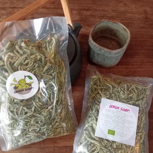 Puede incluir: Dos bolsas de plástico transparente llenas de hojas verdes secas, etiquetadas "Lemon Soup!" con un gráfico de limón. También hay una tetera pequeña y oscura con un asa de madera y una taza de cerámica verde.