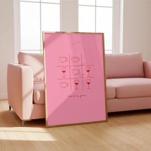 May include: A framed pink print depicting a tic-tac-toe game with coffee cups and wine glasses. The words "the daily game" are printed at the bottom. The print is in a gold frame, leaning against a wall.