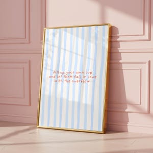 Könnte beinhalten: Ein gerahmter Kunstdruck mit einem goldenen Rahmen. Das Kunstwerk zeigt einen hellblau-weiß gestreiften Hintergrund mit dem Text "fill up your own cup and let them fall in love with the overflow" in roter Schrift.