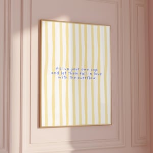 Könnte beinhalten: Ein gerahmter Kunstdruck mit gelb-weiß gestreiftem Hintergrund. Der Druck zeigt blauen Text mit der Aufschrift "fill up your own cup and let them fall in love with the overflow". Der Rahmen ist hellbraun.