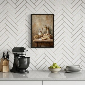 May include: A framed still life painting of bread, flour, and wheat on a wooden table. A black stand mixer, a knife block with knives, a bowl of green apples, and a stack of plates are on the counter below the painting.