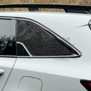 May include: White SUV with black accents, including wheels and roof rack. The rear window features a black and white topographic design. Red taillights are visible. The vehicle is parked on a gravel surface.