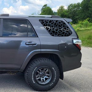 May include: A gray SUV with a black leopard print window decal. The vehicle has black tires and a silver roof rack. The background includes a cloudy sky and green trees.