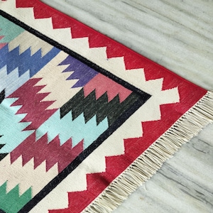 May include: A woven rug featuring a geometric pattern of interlocking zig-zag shapes in shades of red, blue, purple, green, and cream. The rug is framed by a bold red border with a white serrated edge and finished with cream-colored fringe.