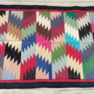 May include: A rectangular woven rug featuring a repeating vertical zigzag pattern in vibrant colors including red, blue, green, black, and beige. The rug is framed by a dark blue border and finished with white fringe tassels on the two shorter ends.