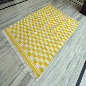 Op de afbeelding: Een rechthoekig tapijt met een herhalend geel en wit pijlenpatroon. Het tapijt heeft een witte franje aan de korte uiteinden en een witte rand. Het is gemaakt van geweven materiaal en ligt op een marmeren vloer.