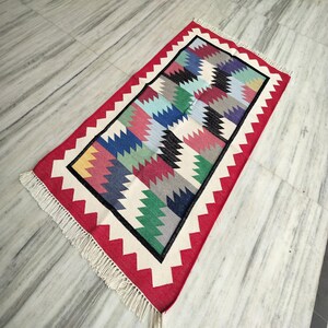 Puede incluir: Alfombra rectangular tejida a mano con un vibrante patrón geométrico. La alfombra presenta un borde rojo con zigzags blancos, un borde interior negro y blanco, y un diseño central de zigzags coloridos en varios tonos. La alfombra tiene flecos en los extremos cortos.