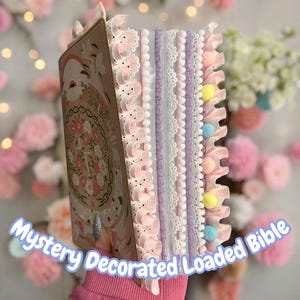 May include: A decorative Bible with a floral design on the cover. The edges are adorned with pink, white, and lavender lace, ruffles, and pom-poms. The text "Mystery Decorated Loaded Bible" is visible.