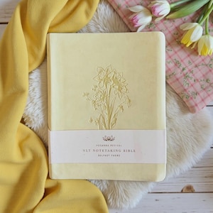 May include: A pale yellow hardcover Bible featuring an embossed floral design on the front cover. A white paper band wraps around the center, printed with the text: HOSANNA REVIVAL, NLT NOTETAKING BIBLE, BELFAST THEME, and a small floral icon.