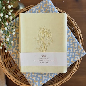 May include: A light yellow NLT Notetaking Bible with embossed floral design. The book is on a patterned cloth inside a woven basket. The text on the book reads "Hosanna Revival" and "Belfast Theme".