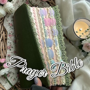 May include: A green leather-bound Prayer Bible with decorative lace edging and pastel-colored page tabs. The tabs are in shades of pink, blue, and cream. The words "Prayer Bible" are written in a cursive font.