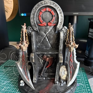 May include: A grey and black 3D printed throne sculpture featuring a circular backrest with engraved runes and a red Omega symbol. Two large silver blades with gold chains flank the seat, while a small red axe and a skull rest at the base.