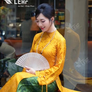May include: A woman in a traditional Vietnamese Áo Dài dress. The dress is a bright yellow with a floral pattern, combined with green trousers. She holds a white, intricately carved fan. The image includes the "LEAF" logo and text.