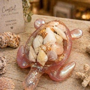 May include: A decorative, pink, glittery resin turtle figurine filled with various seashells. The turtle has a pearlescent sheen and is displayed on a wooden surface. A tag that reads "Coastal Collection" is visible in the background.