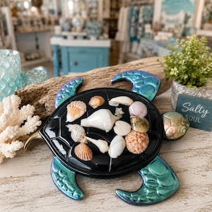May include: A decorative turtle figurine featuring a glossy black shell adorned with various small, natural seashells. The turtle has metallic teal-green flippers and a head, with a textured surface. The shell is covered in white, tan, and pink shells.