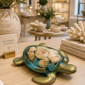May include: A decorative turtle figurine with a blue and gold finish, filled with seashells. The figurine is part of the "Ocean Breeze Collection" and sits on a wooden surface. Books titled "Coastal Living" and "The Kinfolk Home" are nearby.