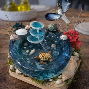 May include: A miniature decorative fountain with a blue resin basin, featuring a small turtle figurine and cascading water. The fountain is adorned with seashells, moss, and a butterfly accent. It sits on a wooden base.