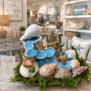May include: A decorative tabletop water fountain featuring a large white conch shell at the top, cascading water over blue resin pools, surrounded by various natural seashells, green moss, and a small turtle figurine. A sign reads Coastal Collection.