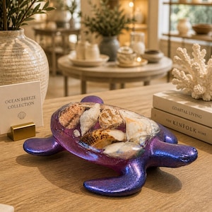 May include: A decorative turtle figurine with a shimmering purple body and a clear shell filled with seashells. The figurine is on a wooden surface, with books and other decor in the background. The sign reads "Ocean Breeze Collection."