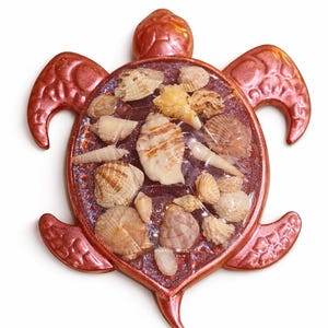 May include: A decorative turtle-shaped object with a metallic rose-colored finish. The turtle's shell is filled with various seashells. The design includes a head, four flippers, and a tail, all in the same rose color.