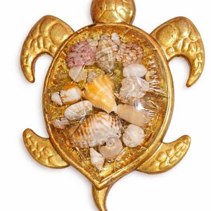 May include: A gold-colored turtle-shaped decorative item. The turtle's shell is filled with a variety of seashells in shades of white, orange, and pink. The turtle's body and flippers have a textured, metallic finish. The item is likely a decorative piece for a coastal or beach-themed setting.