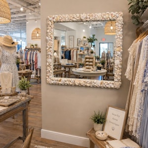 May include: A square wall mirror featuring a frame densely decorated with various white and cream-colored seashells. Below the mirror sits a wooden sign in a frame with the text beachy keen boutique and a small seashell icon printed on it.