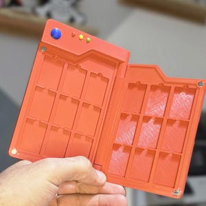 Könnte beinhalten: Offene orangefarbene Kunststoffhülle mit mehreren rechteckigen Fächern, die in einer Hand gehalten wird. Die Hülle hat ein Scharnierdesign und eine Reihe kleiner Knöpfe auf der Oberseite. Der Hintergrund umfasst verschwommene Dekorationsgegenstände auf Regalen.