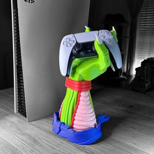 May include: A white and black PlayStation 5 console stands next to a colorful controller stand. The stand features a green hand holding a white controller, with a red, pink, and blue base. The controller is white and black.