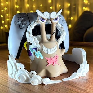 Op de afbeelding: Een bruin en wit cartoon slak beeldje met een wit wolkenontwerp. De slak heeft paarse ogen en een roze ster op zijn lichaam. Achter de slak staat een witte en zwarte gamecontroller.