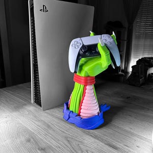 Puede incluir: Una consola PlayStation 5 blanca colocada verticalmente junto a un soporte decorativo para mando con forma de mano verde con garras y uñas blancas, que lleva una muñequera roja, un antebrazo rosa y una base azul, sosteniendo un mando blanco y negro.