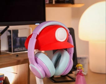 May include: Red and white novelty headphone stand shaped like a video game character's hat, holding lavender and light blue headphones. The stand is on a desk with a Princess Peach figurine and a lamp.