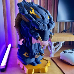 Peut inclure: Un buste de créature bleu foncé et doré présentant une tête texturée et blindée avec des crocs acérés, un museau rose clair et des détails de cou segmentés marron. La figurine est montée sur une base hexagonale dorée aux motifs sculptés.