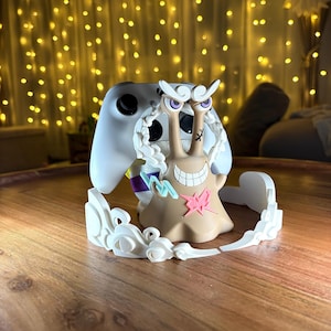 Op de afbeelding: Een beige, slakvormige controllerstandaard met een brede grijns, paarse ogen en een roze bliksemembleem op de borst. Het figuur is omringd door witte, wolkachtige accenten en houdt een witte gamecontroller achter het hoofd vast.
