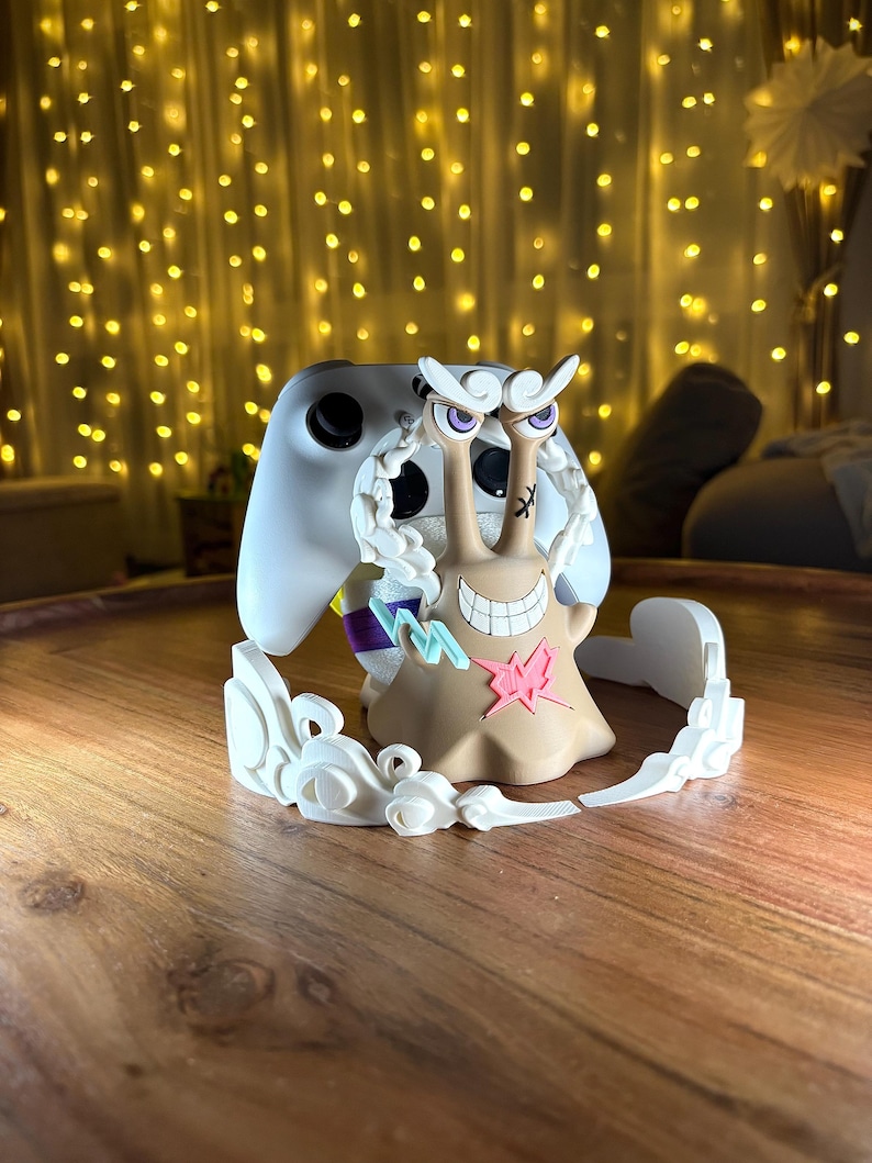 Op de afbeelding: Een beige, slakvormige controllerstandaard met een brede grijns, paarse ogen en een roze bliksemembleem op de borst. Het figuur is omringd door witte, wolkachtige accenten en houdt een witte gamecontroller achter het hoofd vast.