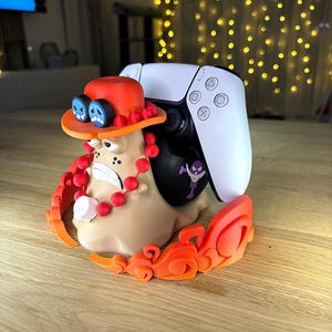 Op de afbeelding: Een beige slakvormige controllerstandaard met een figuur met een oranje hoed, blauwe bril en rode kralen, die een wit-zwarte gamecontroller vasthoudt. De basis is versierd met oranje vlamachtige accenten en een kleine paarse schedelsticker.