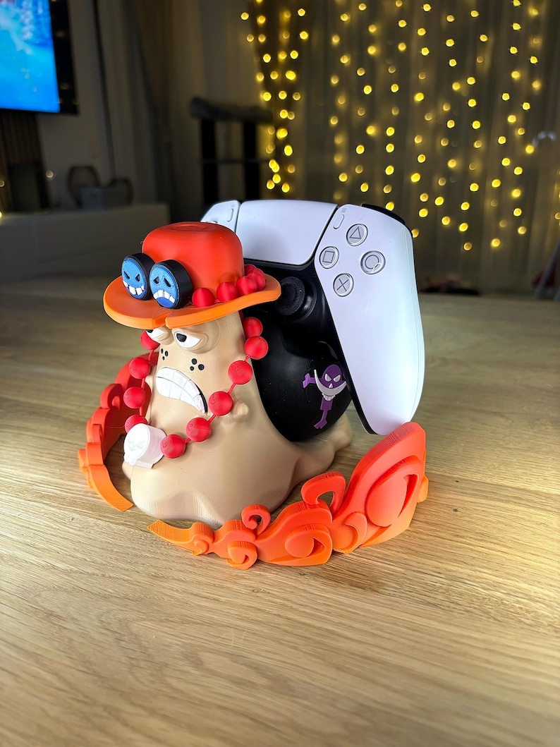Op de afbeelding: Een beige slakvormige controllerstandaard met een figuur met een oranje hoed, blauwe bril en rode kralen, die een wit-zwarte gamecontroller vasthoudt. De basis is versierd met oranje vlamachtige accenten en een kleine paarse schedelsticker.