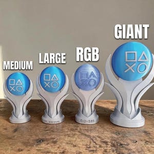 Op de afbeelding: Vier trofee-achtige standaards met blauwe bolvormige toppen met in reliëf PlayStation-controllersymbolen. De standaards zijn gelabeld MEDIUM, LARGE, RGB en GIANT. Elke basis is zilverkleurig en heeft een gebogen, vlamachtig ontwerp.