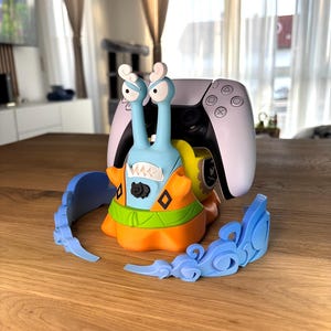 Op de afbeelding: Een lichtblauwe en oranje controllerstandaard in de vorm van een slak met een wit-zwarte gamecontroller op de rug. De slak heeft twee hoge oogstelen, een brede mond met scherpe tanden en wordt omringd door twee losse, lichtblauwe golfvormige onderdelen.