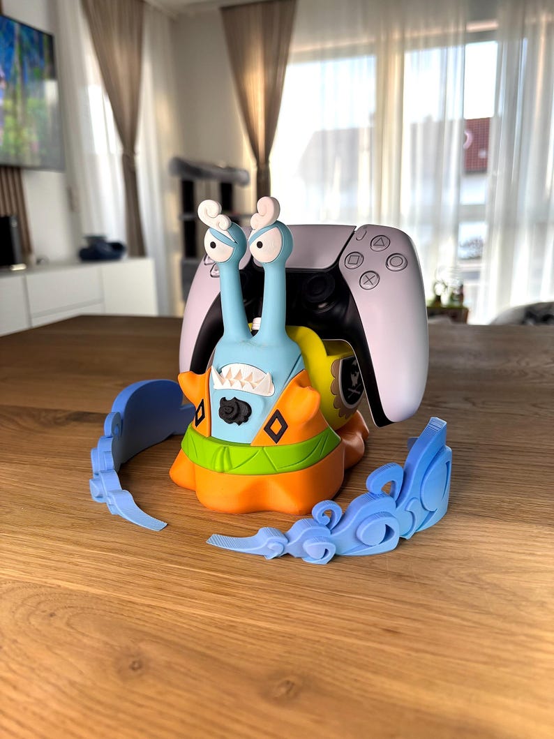 Op de afbeelding: Een lichtblauwe en oranje controllerstandaard in de vorm van een slak met een wit-zwarte gamecontroller op de rug. De slak heeft twee hoge oogstelen, een brede mond met scherpe tanden en wordt omringd door twee losse, lichtblauwe golfvormige onderdelen.