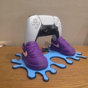 Op de afbeelding: Een wit en zwart videogame-controller die in twee paarse sneakers met lichtroze swoosh-logo's rust. De sneakers zijn bevestigd op een platte, lichtblauwe basis in de vorm van een vloeistofspat met een donkerblauwe rand, als een standaard.